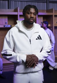 Haut de sweatshirt blanc zippé avec un tissu texturé et des accents noirs, associé à un pantalon assorti. Les caractéristiques notables incluent un col haut et des poches.