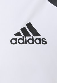Logo Adidas noir avec trois bandes inclinées au-dessus du nom de la marque sur un fond en tissu blanc.