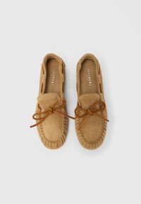 Mocassins en daim marron clair avec un bout rond, comprenant des détails de couture et un accent de lacet noué à l'avant. Nom de la marque visible à l'intérieur.