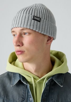 Joven con un gorro de punto gris, sudadera con capucha verde clara y chaqueta vaquera, mirando hacia su izquierda sobre un fondo liso.