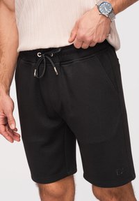 Homme portant un short noir à cordon et une chemise beige côtelée, ajustant l'ourlet de la chemise, avec une montre-bracelet argentée au poignet gauche.