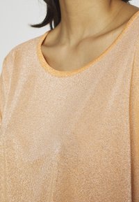 Välkande persikofärgad topp gjord av ett texturerat tyg, med en rund halsringning som har en ljus orange kant. Subtil lyster förbättrar designen.