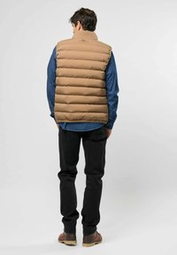 Béžová péřová vesta s horizontálním prošíváním, vysokým límcem a značkovou štítkem, nošená přes džínovou košili, doplněná černými džíny.