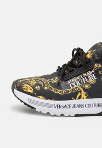 Svart sneaker med guldkedjemönster, övre del i tyg, gummisulor och logotypen "VERSACE JEANS COUTURE" tryckt i vitt på sidan.