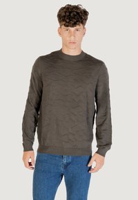 Dunkelgrüner Pullover mit strukturiertem Wellenmuster, rundem Halsausschnitt und gerippten Bündchen und Saum. Kombiniert mit blauen Jeans.