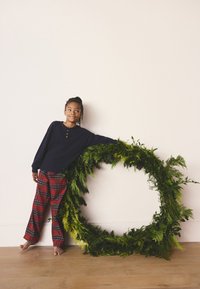 Child in navy long-sleeve shirt with wooden buttons and red plaid pajama pants, leaning against a circular green wreath of foliage.