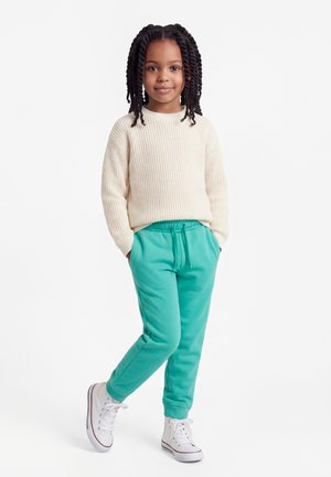Mladá dívka s copánky, oblečená v krémovém pleteném svetru, tyrkysových teplácích a bílých vysokých teniskách, stojící s rukama v kapsách.