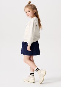 Grauer Sweatshirt, marineblauem Rock mit Tasche, cremefarbene Sneaker und gestreifte Socken. Kind steht seitlich, Haare zu einem Dutt gebunden, vor einem neutralen Hintergrund.