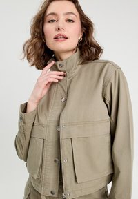 Chaqueta caqui con un cuello alto, que cuenta con dos bolsillos frontales, herrajes plateados y cierre de cremallera. Tejido duradero con una superficie texturizada.