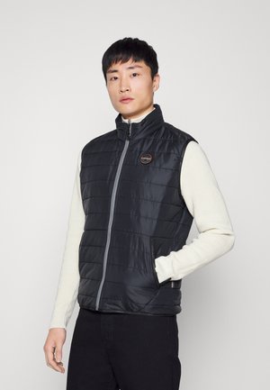 Black puffer vest with zip closure, vertical quilted pattern, high neck, side pockets, and a logo patch on the chest.