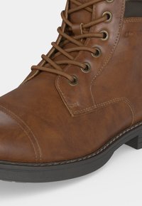 Botte en cuir marron à lacets avec œillets métalliques, coutures détaillées et semelle en caoutchouc noir visible sur fond blanc.