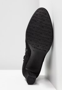 Semelle d'une botte pointue en daim noir montrant le motif de la bande de roulement « Tamaris » et la taille 37, talon marqué « ANTI SHOK » sur fond de sol et mur blanc.