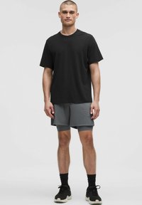 Un homme se tient avec confiance dans un t-shirt noir et un short de sport gris, portant des chaussettes et des chaussures noires, devant un fond clair et uni.