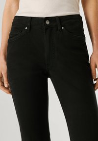 Femme portant un jean noir taille haute avec poches avant et fermeture à boutons, associé à un haut sans manches côtelé blanc.