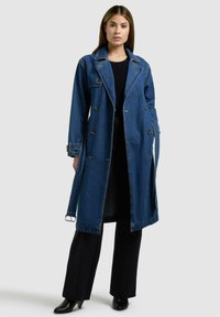 Kvinde iført en lang blå denim trenchcoat over sorte bukser og sorte støvler med hæl, stående foran en ensfarvet lys baggrund.