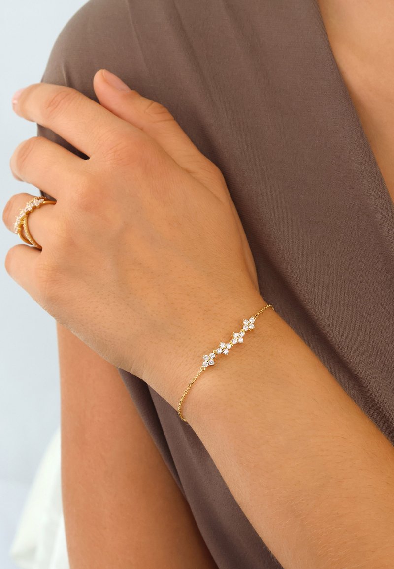 Gold bracelet with five clear crystal flowers, featuring delicate chain links. Hand wearing the bracelet rests on a brown fabric surface.