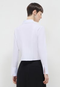 Camicia bianca con colletto, maniche lunghe, silhouette aderente e spacchetti laterali ai polsini, abbinata a una gonna nera. Tessuto dalla texture liscia.