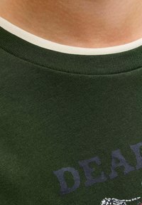 Gros plan sur un t-shirt à col côtelé vert superposé à une chemise blanche, montrant partiellement un texte bleu foncé et un petit design graphique.