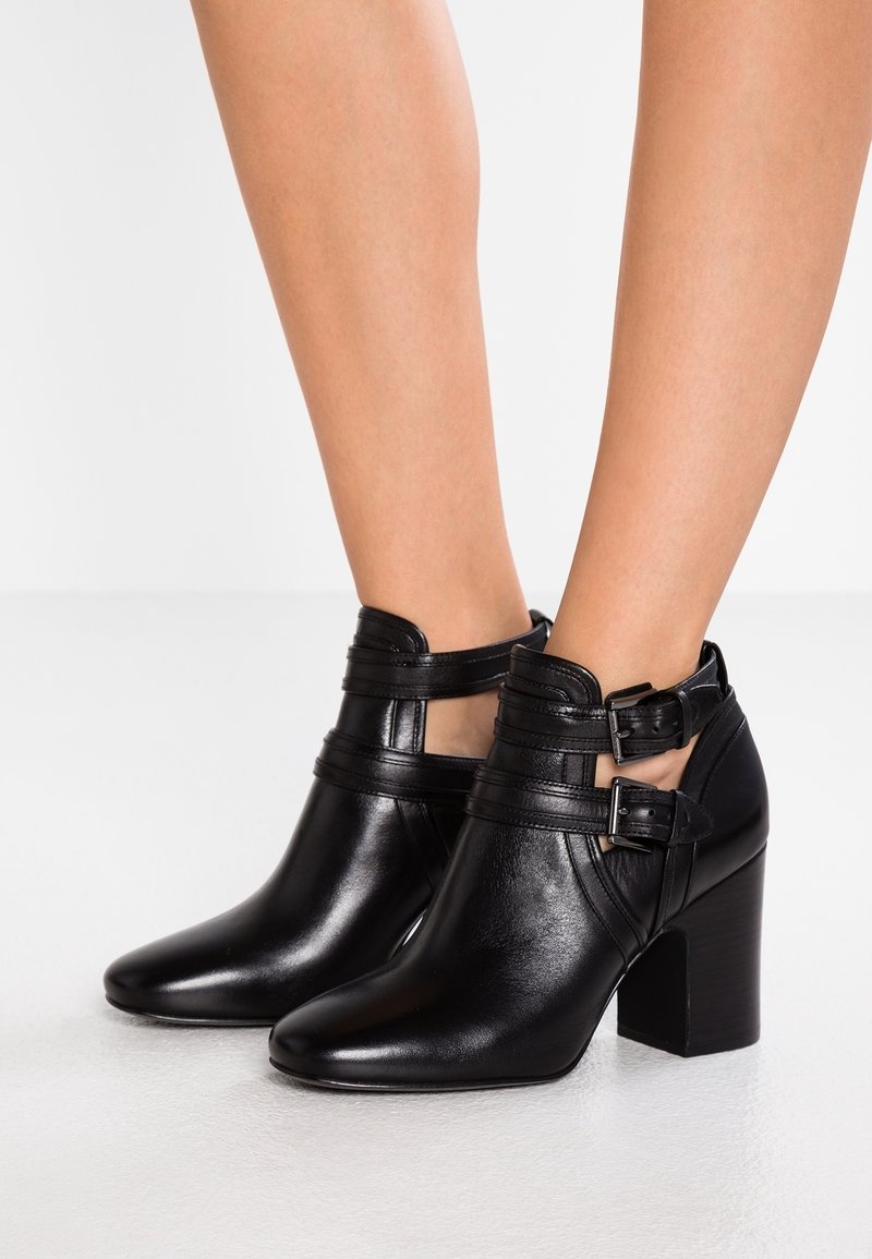Bottines en cuir noir avec deux brides à boucle et talons blocs portées sur des jambes nues sur un fond blanc.