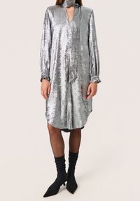 Vestido metálico plateado con escote en V profundo, mangas largas y cuello tipo lazo. Tiene un corte holgado y un dobladillo redondeado. Lleva puestas medias oscuras y tacones.