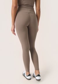 Simított, középbarna anyagú leggings, testhez simuló fazonban, magas derékkal, fehér sportcipővel, fekete részletekkel párosítva.