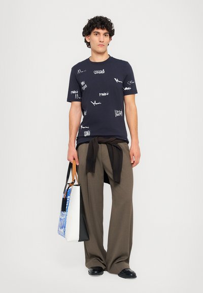 Marineblå bomulds-t-shirt med hvide tekstmønstre, parret med brune vidde bukser, mens der holdes en to-farvet tote bag med et blåt design.