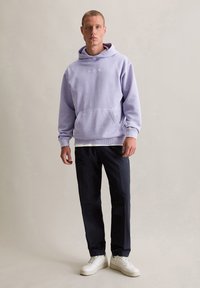 Felpa lilla con cappuccio e tasca a marsupio, polsini e orlo a coste, abbinata a pantaloni scuri e sneaker bianche, in piedi su uno sfondo neutro.