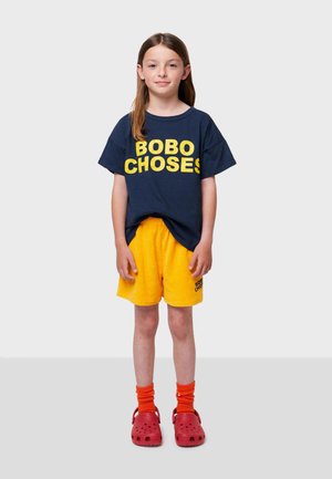 T-shirt marine avec un texte jaune vif "BOBO CHOSE", associé à un short jaune. Les accessoires incluent des crocs rouges et des chaussettes orange.