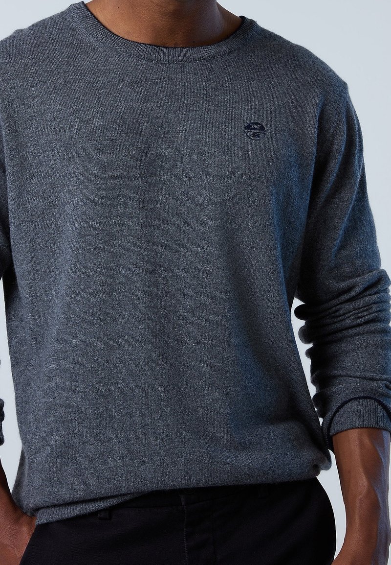 Homme portant un pull uni gris foncé à manches longues avec un petit logo circulaire noir sur la poitrine gauche, associé à un pantalon noir.