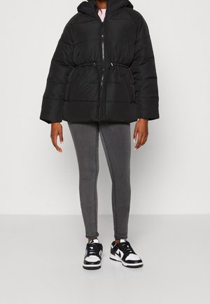 Person trägt eine schwarze Steppjacke mit Frontreißverschluss und Kordelzug in der Taille, graue Skinny-Jeans und schwarz-weiße Sneakers.