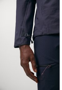 Navy waterproof jacket featuring adjustable cuffs, a textured finish, and a side zip pocket with contrasting pull cord and logo detail.