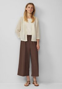 Eine Frau trägt eine beigefarbene Netzstrickjacke über einem cremefarbenen Oberteil, braune weit geschnittene, knöchellange Hosen und Sandalen mit Leopardenmuster, während sie vor einem grauen Hintergrund steht.
