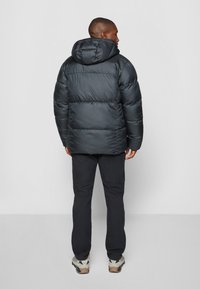 Chaqueta acolchada en gris oscuro con capucha, que presenta secciones acolchadas y una textura suave. Combinada con pantalones negros y zapatillas de senderismo grises.