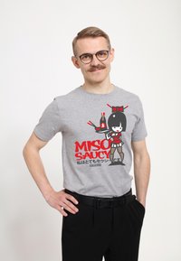 Camiseta gris de manga corta con un gráfico colorido de un personaje sosteniendo una botella, con el texto "MISO SAUCY" en letras rojas en negrita.