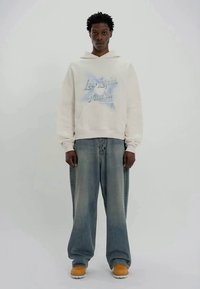 Homem a usar um hoodie branco com texto azul, jeans largos e desbotados, e botas amarelas, em pé contra um fundo claro e liso.
