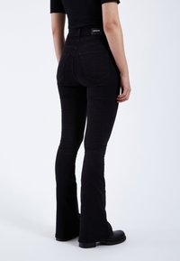 Jeans flared de talle alto en negro con una textura suave, dos bolsillos traseros y una etiqueta minimalista en la cintura.