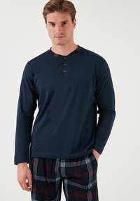 Camiseta de manga larga azul marino con un cuello Henley abotonado y pantalones de pijama de cuadros que presentan un diseño de cuadros rojos y blancos sobre un fondo oscuro.