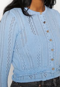 Suéter de punto azul cielo con textura acanalada, frente con botones y patrones de ojetes. Diseño cropped con detalle de dobladillo fruncido.