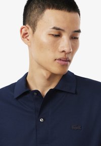 Marinblå pikétröja med korta ärmar, klassisk krage, två knappslitsar och en svart broderad logotyp på bröstet. Slät tygtextur.