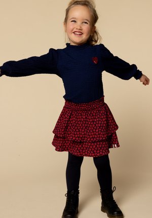 Langarmige marineblauwe top van textiel met een hartdetail, gecombineerd met een rode gelaagde rok met een bloemenpatroon en zwarte enkellaarsjes.