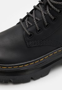 Botín de cuero negro con cordones grises, suela de goma resistente, costuras amarillas, ojales reforzados y un acabado suave.