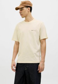 Light beige cotton t-shirt with short sleeves, a crew neckline, and a small logo on the left chest. Paired with dark jeans.