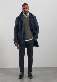 Navy hooded coat with snap closures, layered over a textured grey sweater and white shirt, paired with navy pants and black shoes.