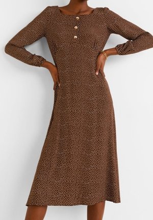 Braunes Midikleid mit kleinen beigen Punkten, langen Ärmeln, quadratischem Ausschnitt, drei Knöpfen auf der Brust, getragen mit Händen in der Hüfte.