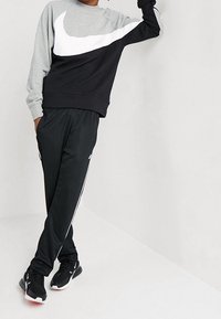 Person klädd i en svart och grå färgblockerat Nike-tröja, svarta Nike-träningsbyxor med vita sidostripes och svarta Nike-sneakers.