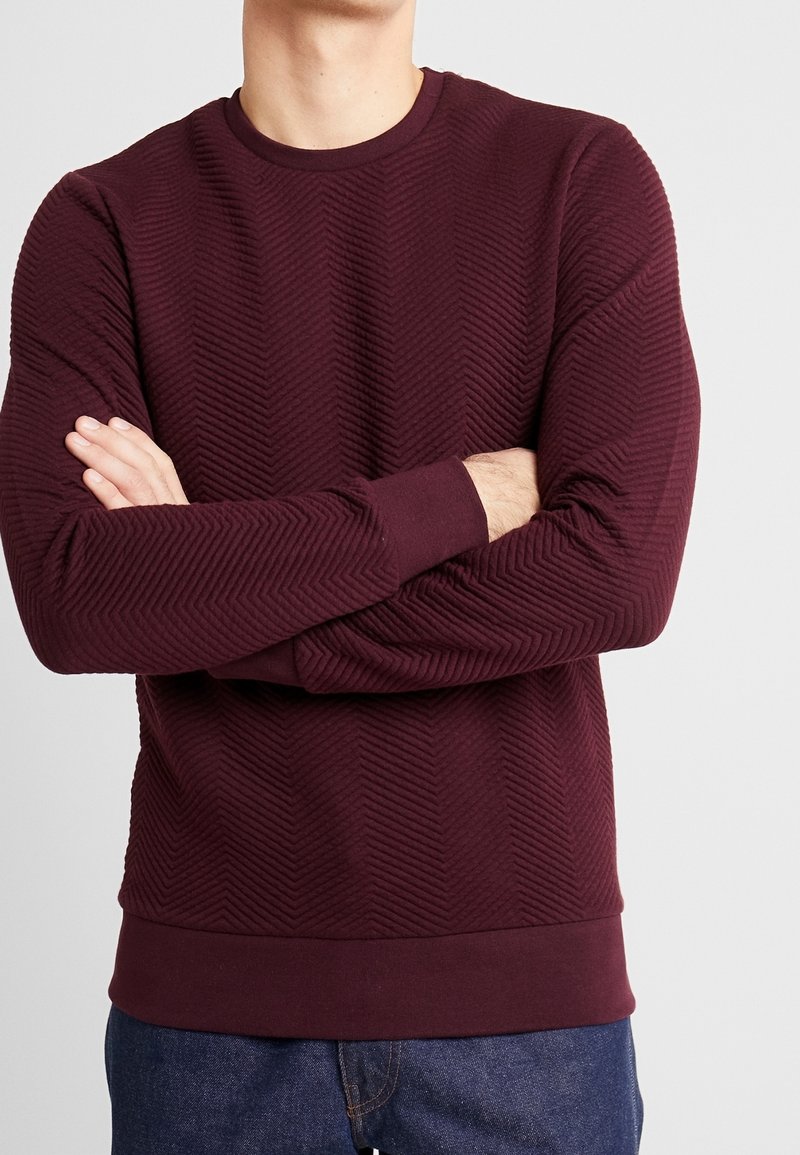 Uomo che indossa un maglione bordeaux a trama con le braccia incrociate, abbinato a jeans blu scuro, su uno sfondo chiaro e uniforme.