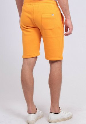 Homme portant un short orange vif arrivant aux genoux avec une poche arrière et des baskets blanches, debout devant un fond uni.