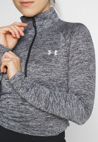 Harmaa, teksturoitu neljännesvetoketjullinen yläosa, jossa korkea kaulus. Siinä on pitkät hihat ja valkoinen Under Armour -logo rinnassa.
