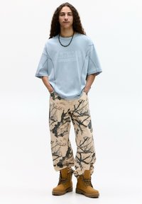 Světle modré volné triko s potiskem, vzorované béžové kalhoty s černými větvemi a hnědé šněrovací boty se silnou podrážkou.