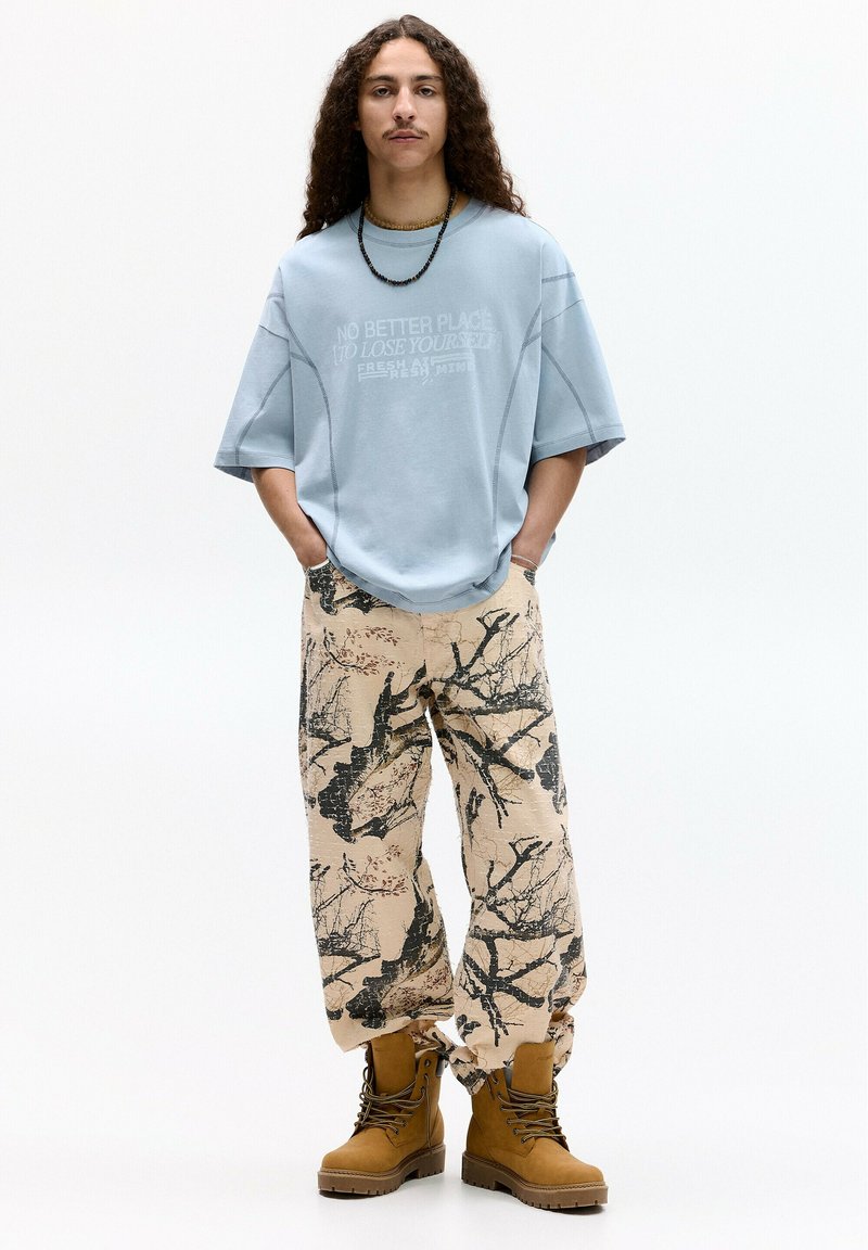 Světle modré volné triko s potiskem, vzorované béžové kalhoty s černými větvemi a hnědé šněrovací boty se silnou podrážkou.